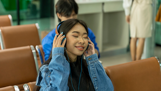 Young girl enjoys listening music with wireless headset while waiting at an airport lounge before boarding. Female traveler feels happy with music waiting for her trip at a train terminal
