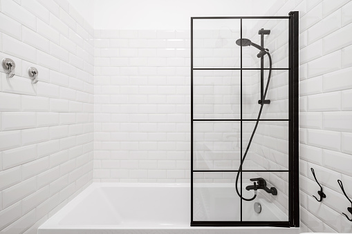 stylish bathtub, black faucet, shower nozzle head and glass partition in bathroom with white tile, contemporary interior