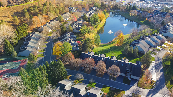 Aerial view lush green apartment complex in nature settings with lake water fountain, tennis court in suburban Rochester, Upstate New York, USA. Row of rental townhome condos colorful autumn leaves