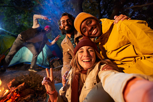 Mobile selfie of Multiracial smiling friends in the forest camping. Young adults with traveling spirit spending time in the countryside at night. Group of happy cheerful colleagues outdoors.