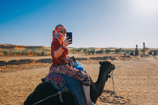 asian Chinese female tourists riding dromedary camel train crossing Sahara Desert Morocco