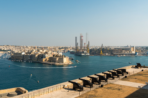 Magnificent view of the sea harbor and the old fortress on the other side of the bay, medieval Maltese architecture, yellow local stone, ships in the parking lot, huge cranes of the cargo port, ancient English cannons, panoramic view, blue sea and sky, urban seascape, travel and leisure , summer, Europe.