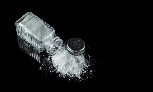 Photo of Heap of salt from a salt shaker on black background. The concept of excessive salt intake that can be the cause of diabetes or other diseases