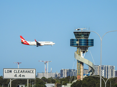 The air traffic control tower at Sydney Kingsford-Smith Airport.  The curved structure is an emergency exit.  In the distance is a Qantas 737-838, VH-VZS, flight number QF539 from Brisbane, coming into land.  The road sign refers to the tunnel under the airport runway.  This image was taken from Kyeemagh Avenue, Kyeemagh on a sunny afternoon on 5 February 2023.