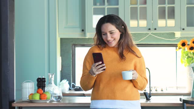 Young woman in the kitchen. She is drinking first coffee while using mobile phone