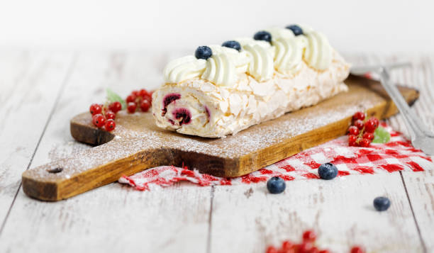 砂糖粉を木の板に詰めたベリーとジャムの白いロールケーキ。白い素朴な背景にホイップクリーム、ブルーベリー、赤スグリで飾られたデザートケーキ