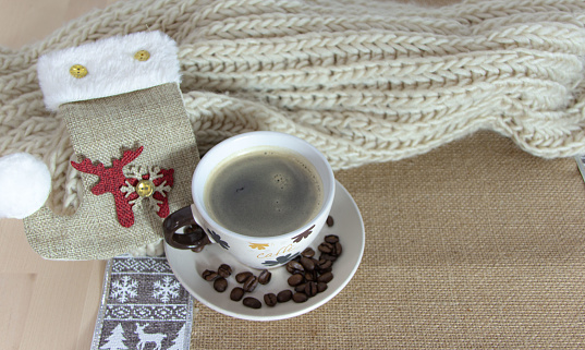 Cup of coffee on a plate with some coffee beans, next to a Christmas stocking over a wooden table and a jute tablecloth. Cozy Christmas decoration.