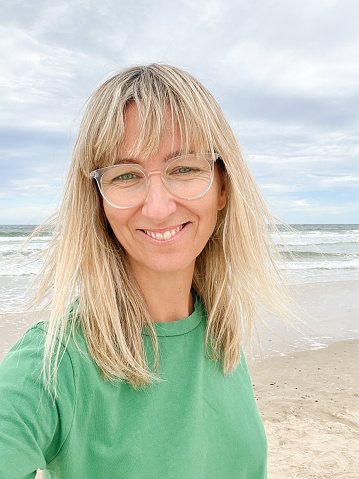 young tourist woman long blond hair taking selfie with smart phone, wearing green casual shirt on a sandy beach in Australia
