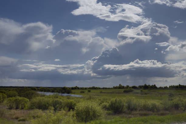 Dramatic sky with blue fluffy thunderstorm clouds and sunbeams in fresh bright spring landscape above green meadow with bushes, pond in motion. Breathtaking weather in rural scene.