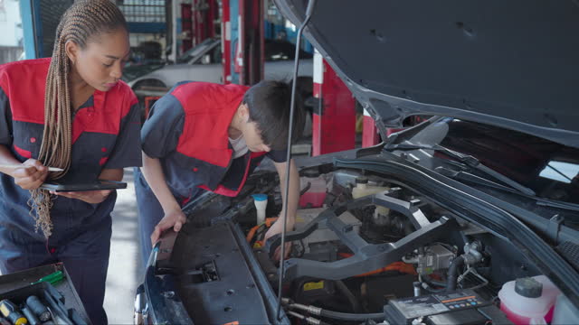 Close-up view of two professional garage mechanics, male and female working with multimeter or voltmeter checking a car battery level while young Asian female writing information to digital tablet. Open electric car's hood with car details. Service center