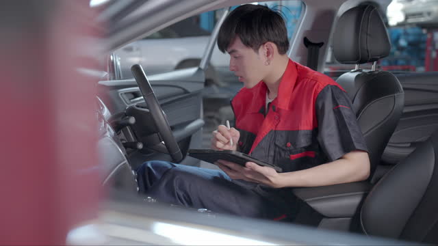 Handsome Asian mechanic engineer using a laptop to work at the repair garage. Professional engineer male sitting inside electric car and using digital tablet for diagnosing car's problem in auto repair service.