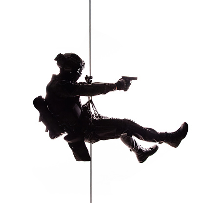 Silhouette of police officer in tactical gear descending from a height, rope exercises with weapons. Tactical rappelling, anti-terror or counter terrorism operation in darkness in rappelling harness, white background