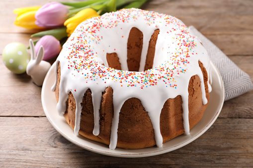 Glazed Easter cake with sprinkles, decorative bunny, painted egg and tulips on wooden table, closeup