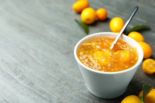 Delicious kumquat jam in bowl and fresh fruits on grey table, space for text