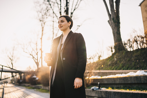 A young, happy businesswoman holding takeaway coffee. A happy businesswoman having a coffee break stock photo. Young business woman having a cup of coffee in a park during a break.