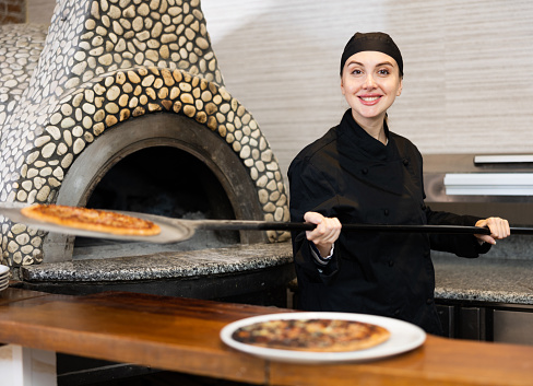 Portrait of smiling pizza chef at work, woman taking pizza from oven at restaurant kitchen