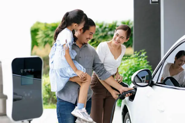 Progressive young parent teach daughter how to recharge or refuel EV car at home charging station. Green and clean energy from electric vehicle for healthy environment. Eco power from renewable source