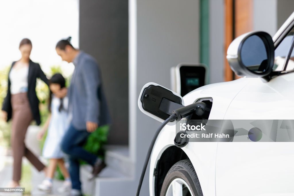 Focus home charging station for EV car, blur progressive family in background. Focus closeup electric vehicle recharging battery from home electric charging station with blurred family in background. Renewable clean energy car for progressive eco awareness lifestyle concept. Electric Car Stock Photo