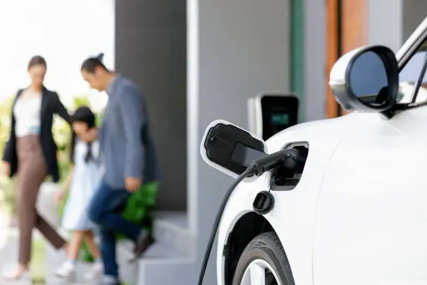 Focus closeup electric vehicle recharging battery from home electric charging station with blurred family in background. Renewable clean energy car for progressive eco awareness lifestyle concept.