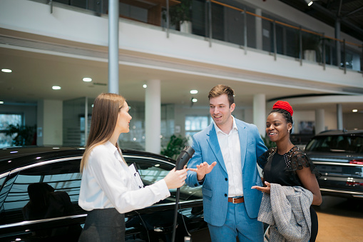 Salesman showing new electric car to multiracial couple at showroom. Female sales manager holding electric car charger and explaining the benefits of electric car to buyer couple at dealership.
