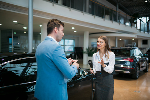 Saleswoman holding car charging adapter and explaining the features of an electric car in showroom. Female sales manager selling electric car to a male customer. Customer discussing with saleswoman at electric car dealership.