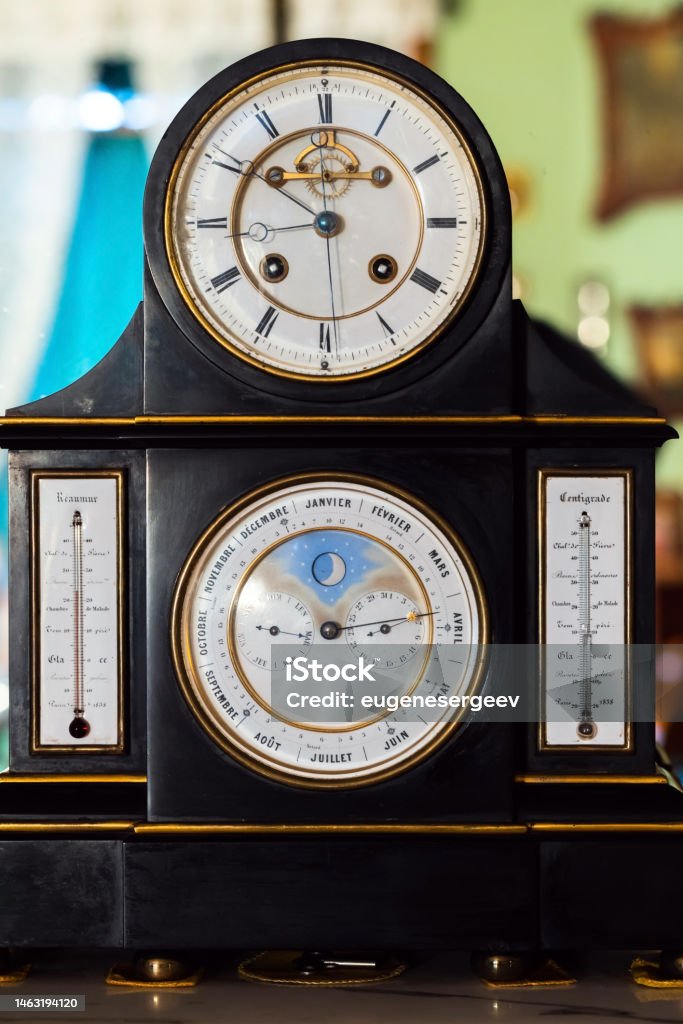 Vintage table clock with white deal and retrograde calendar Vintage table clock with white deal, retrograde calendar, moon phase indicator, Reaumur and Celsius temperature scales. Close up frontal photo Antique Stock Photo