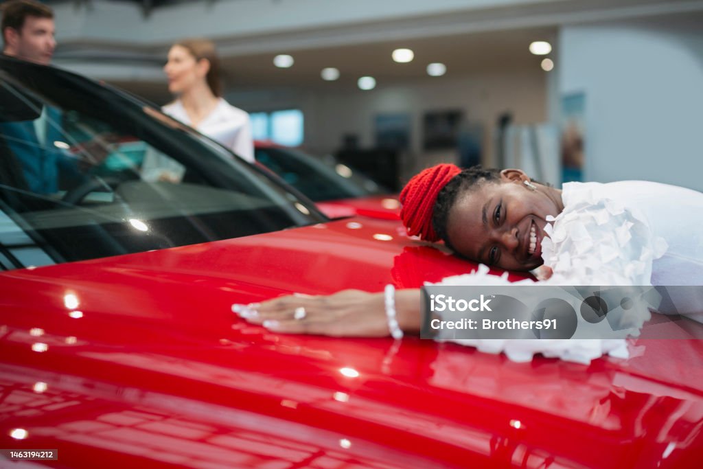 Happy young woman giving her new car a big hug Happy young woman giving her new car a big hug and smiling on showroom. African female feeling happy after buying a new car at auto dealership. Humor Stock Photo