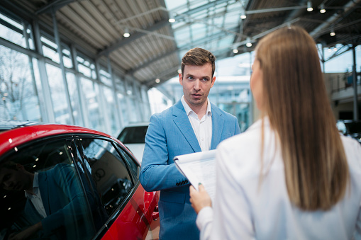 Young man having conversation with female sales assistant at luxury auto showroom. Customer talking with sales assistant while buying new car.