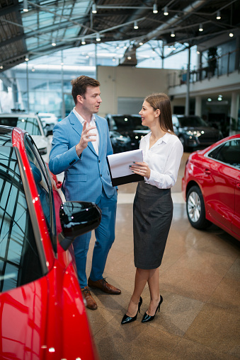Male customer talking with saleswoman holding some documents at car showroom. Businessman buying a new car at auto dealership meeting with sales manager.