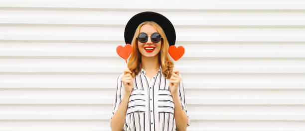 Portrait of beautiful young woman with red heart shaped lollipop wearing sunglasses, black round hat on white background stock photo