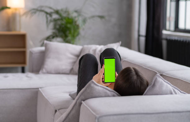 istock Feminine Hand Scrolling Feed on Smartphone with Green Screen Mock Up Display. Female is Relaxing on Sofa at Home, Watching Videos and Reading Social Media Posts on Mobile Device. 1463206929