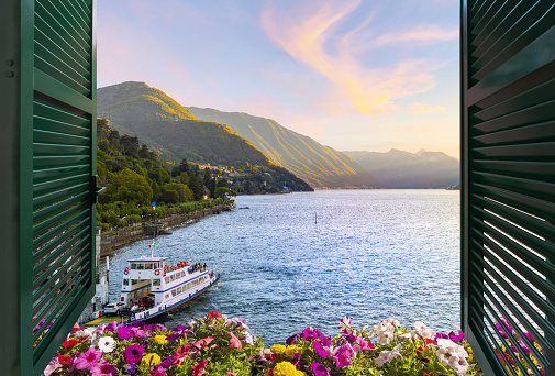 View from a balcony terrace with flowers and shutters of the mountains and lakefront promenade on Lake Como at the town of Bellagio, Italy, under a colorful sunset as a ferry boat sets off from the dock.