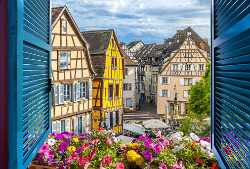 View from an open window overlooking a small square with cafes and half timber frame buildings in the medieval historic center of Colmar, France. Colmar is one of the stops on the Alsatian wine route through Eastern France.