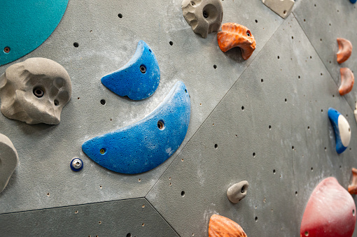 Close up of climbing wall holds and grips of varied shapes and sizes on grey indoor climbing wall at activity centre gym. Blue, red, orange and grey colours.