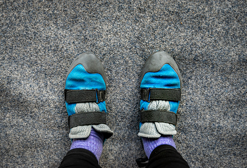 Close up of feet wearing blue and black indoor climbing wall shoes standing on grey soft carpet flooring safety mat. Looking down from above.