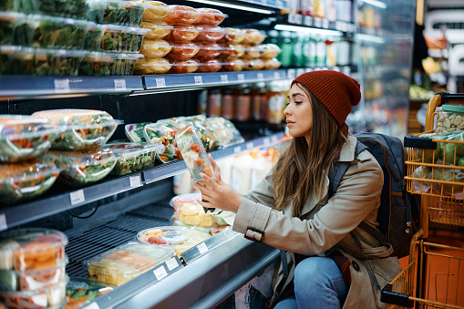 Young woman checking nutrition label on package while buying salad in the supermarket.