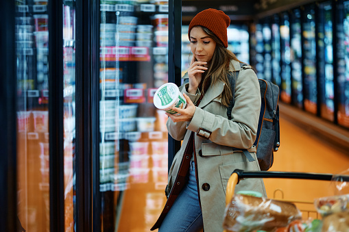 Young woman reading nutrition label while buying dairy product in supermarket.