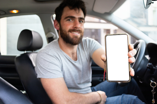 interior shot of a good looking young man working as a driver showing white screen of his phone and smiling