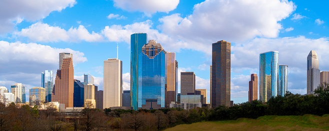 Horizontal, color, Skyline image of Houston, Texas seen across city park with two cyclists (a male and female) riding on a concrete path away from the city.  Backdrop of a beautiful clear blue sky.  Long shadows from trees are present in the lower right foreground in this late afternoon shot.