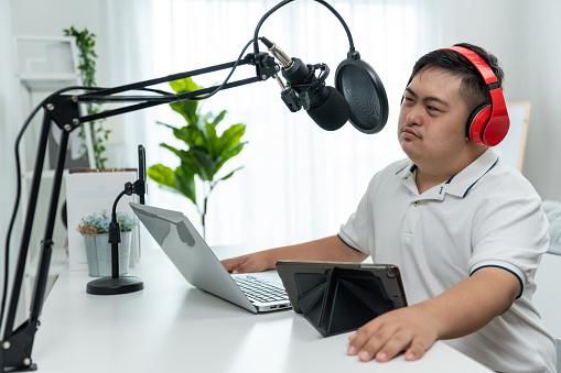 Young down syndrome DJ man wearing headphone and speak on microphone at studio. Teenager patient person feeling happy and relax while recording news podcast show for radio broadcasting at home studio.