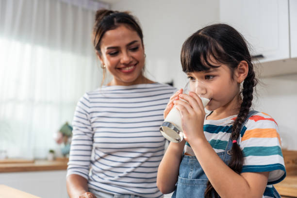 caucasian little kid holding a cup of milk and drinking with mother. attractive mom teach and support young girl daughter take care of her body, sipping a milk after wake up for health care in house. - milk child drinking little girls imagens e fotografias de stock