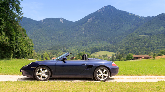 Ruhpolding, Germany - July 24, 2021: Blue roadster Porsche Boxster 986 with Bavarian Alps panorama at German Alpine Road. The car is a mid-engine two-seater sports car manufactured by Porsche.