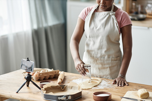 Cropped shot of black woman baking homemade pastry in home kitchen with smartphone camera recording video or livestream, copy space