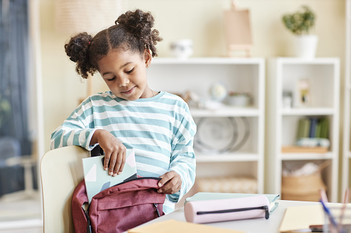 Minimal portrait of cute black girl packing backpack for school in pastel home interior, copy space