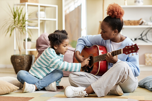 Full length side view portrait of caring mother playing guitar with daughter at home and teaching little girl