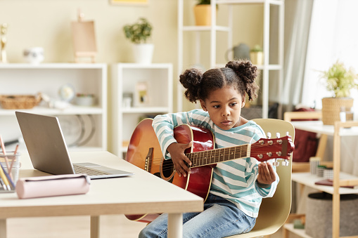 Portrait of black little girl playing guitar at home and watching online music lesson in sunny room , copy space