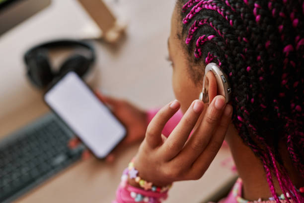 Close up black teenage girl wearing hearing aid on ear Close up of black teenage girl wearing hearing aid on ear and pressing button while using smartphone at home, copy space assistive technology stock pictures, royalty-free photos & images