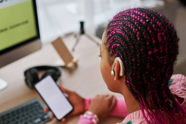 Closeup black teenage girl wearing hearing aids Close up of black teenage girl wearing hearing aid on ear while using smartphone with blank screen at home, copy space assistive technology stock pictures, royalty-free photos & images