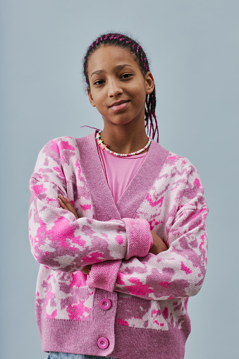 Vertical portrait of black teenage girl wearing pink trendy clothes and posing confidently with arms crossed against blue background