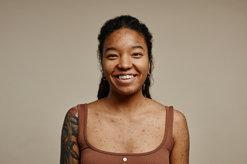 Minimal portrait of young black woman smiling at camera standing against neutral beige background with focus on real skin texture, spots and acne scars